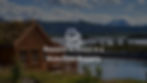 Scenic photograph of a lake house with mountains in the background surrounded by green grass and a lake